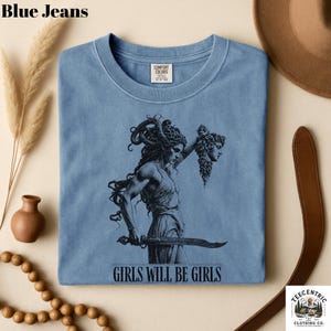 May include: A light blue t-shirt with a graphic of a woman holding a sword and a severed head. The text "GIRLS WILL BE GIRLS" is printed below the graphic. The shirt is made of Comfort Colors material.
