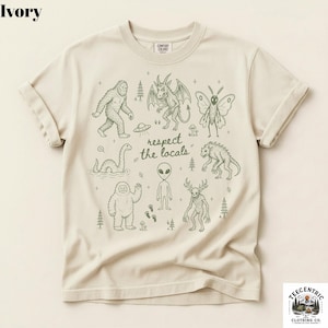 May include: An ivory t-shirt featuring a dark green graphic print of various cryptids and extraterrestrial creatures, including Bigfoot, a mothman, an alien, and a Loch Ness monster, with the text "respect the locals" in the center.