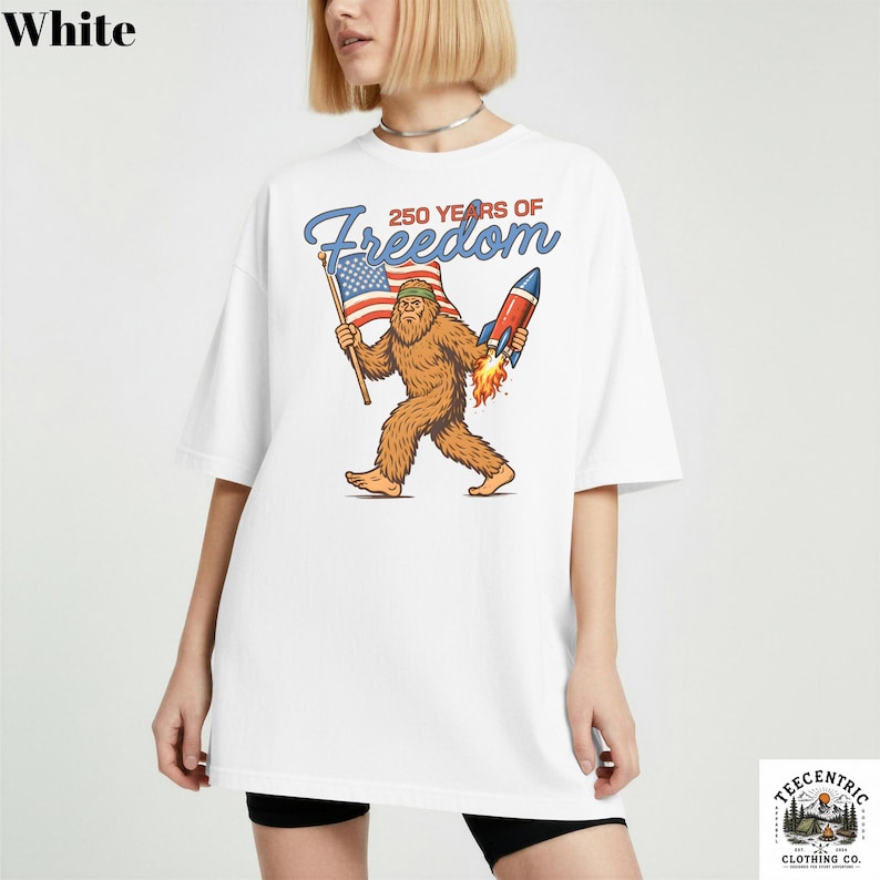 K&ouml;nnte beinhalten: Wei&szlig;es T-Shirt mit einem Cartoon-Bigfoot-Charakter, der eine amerikanische Flagge und eine Rakete mit Flammen h&auml;lt. Der Text "250 YEARS OF Freedom" ist &uuml;ber der Illustration angeordnet. Das Shirt ist von Teecentric Clothing Co.