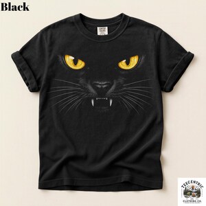 May include: A black short-sleeve t-shirt featuring a graphic print of a cat face with piercing yellow eyes, a dark nose, and visible white fangs. The shirt has a crew neckline and a Comfort Colors brand label printed on the inside of the collar.