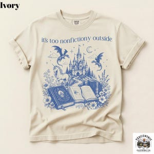 May include: Ivory-colored t-shirt with a blue fantasy graphic. The design features a castle, dragons, a unicorn, books, and the text "it's too nonfictiony outside". The shirt has rolled-up sleeves and a crew neck.