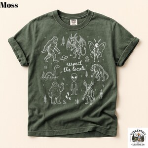 May include: A moss green short-sleeve t-shirt featuring white line art illustrations of various cryptids and an alien, including Bigfoot, the Loch Ness Monster, and a mothman. The text "respect the locals" is printed in the center.