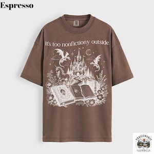 May include: Espresso-colored t-shirt with a fantasy-themed graphic. The design features a castle, dragons, a unicorn, and open books, with the text "it's too nonfictiony outside". The shirt has a crew neck and short sleeves.
