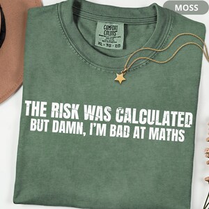 Peut inclure: Un t-shirt vert mousse avec le texte "THE RISK WAS CALCULATED BUT DAMN, I'M BAD AT MATHS" en blanc. Un collier avec un pendentif en forme d'&eacute;toile dor&eacute;e est pos&eacute; sur le t-shirt. Le t-shirt a une &eacute;tiquette Comfort Colors.