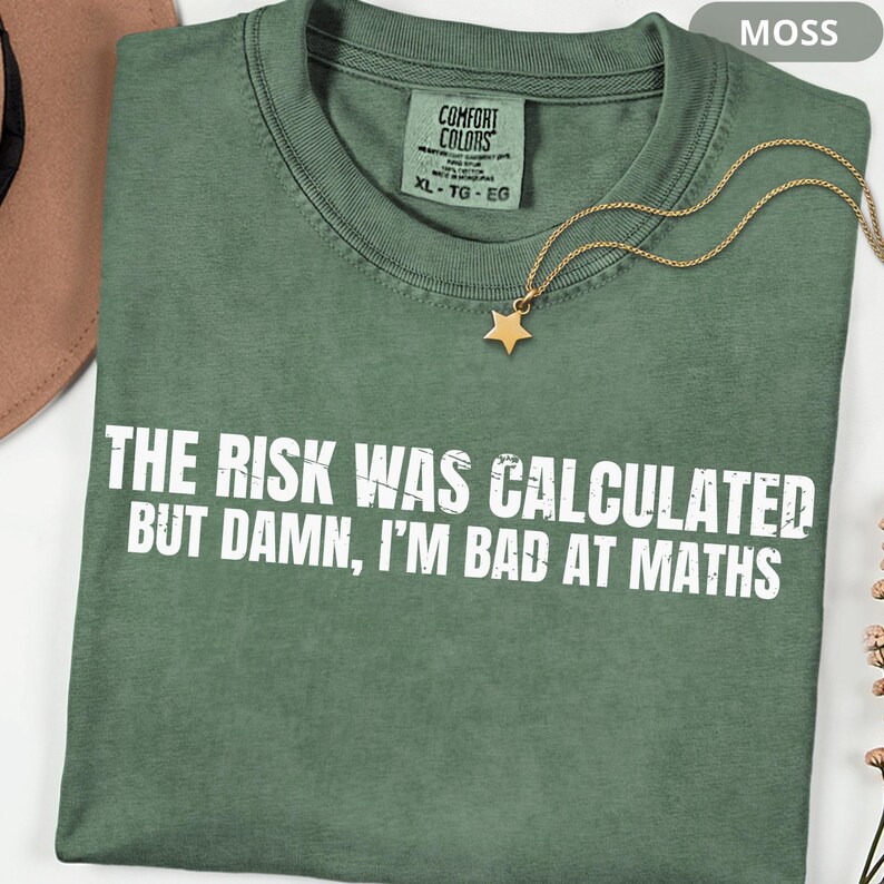 Peut inclure: Un t-shirt vert mousse avec le texte "THE RISK WAS CALCULATED BUT DAMN, I'M BAD AT MATHS" en blanc. Un collier avec un pendentif en forme d'&eacute;toile dor&eacute;e est pos&eacute; sur le t-shirt. Le t-shirt a une &eacute;tiquette Comfort Colors.