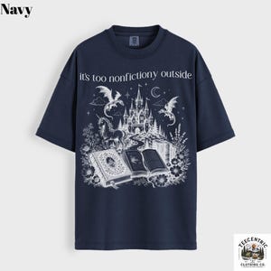 May include: Navy blue t-shirt with a white graphic featuring a castle, dragons, a unicorn, and open books. The text "it's too nonfictiony outside" is printed above the graphic. The shirt has short sleeves and a crew neck.