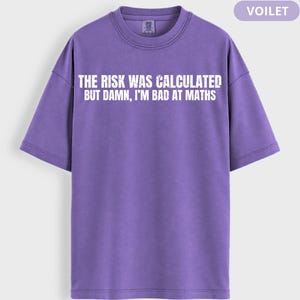 Peut inclure: Un t-shirt violet avec le texte blanc "THE RISK WAS CALCULATED BUT DAMN, I'M BAD AT MATHS". Le t-shirt &agrave; manches courtes a un col rond. Le mot "VOILET" est dans le coin sup&eacute;rieur droit.