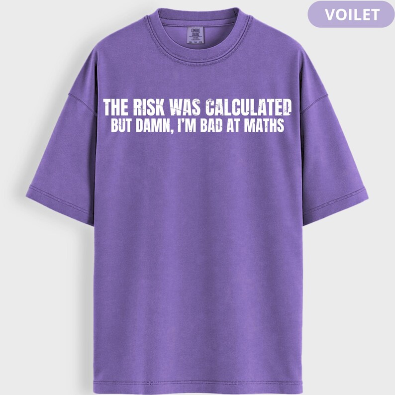 Peut inclure: Un t-shirt violet avec le texte blanc "THE RISK WAS CALCULATED BUT DAMN, I'M BAD AT MATHS". Le t-shirt &agrave; manches courtes a un col rond. Le mot "VOILET" est dans le coin sup&eacute;rieur droit.