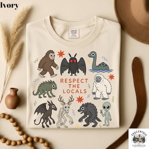 May include: An ivory t-shirt featuring a graphic print of various cryptids and the text "RESPECT THE LOCALS" in orange. The illustrations include a brown Bigfoot, a moth-like creature, a sea monster, a white yeti, a dragon, a deer-like creature, a werewolf, and an alien.