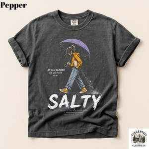 May include: A charcoal gray t-shirt featuring a graphic of a person in a yellow hoodie and blue jeans walking under a purple umbrella in the rain. The text reads All these FLAVORS and you choose to be SALTY, with additional small text below.