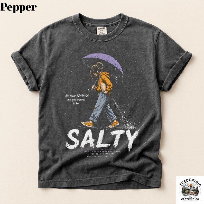 May include: A charcoal gray t-shirt featuring a graphic of a person in a yellow hoodie and blue jeans walking under a purple umbrella in the rain. The text reads All these FLAVORS and you choose to be SALTY, with additional small text below.