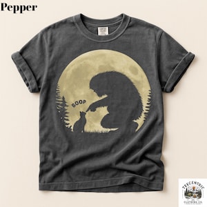 May include: Pepper-colored t-shirt featuring a whimsical graphic of a Bigfoot figure interacting with a cat in front of a full moon. The word "BOOP" is above the cat. The shirt has rolled-up sleeves and a Comfort Colors tag.