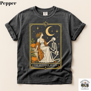 May include: A charcoal gray t-shirt featuring a graphic print of a person in a white dress and an orange shawl sitting with a skeleton under a crescent moon and stars. Two pumpkins are visible. The text THE OCTOBER QUEEN is printed below.