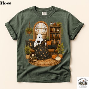 May include: A moss green t-shirt featuring a graphic of a ghost sitting on a couch, reading a book and holding a mug. The scene includes a bookshelf, a black cat, pumpkins, potted plants, and a window with a view of autumn trees.
