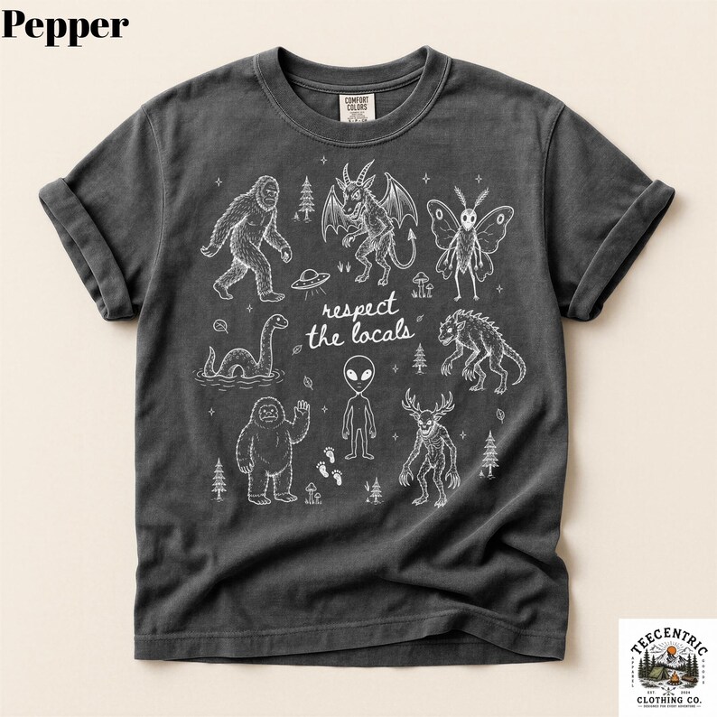 May include: A charcoal gray short-sleeve t-shirt featuring a white graphic print of various cryptids and the text "respect the locals" in the center. The design includes illustrations of Bigfoot, a mothman, an alien, and other creatures.