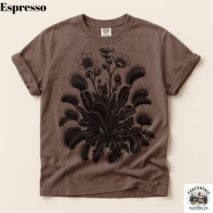 May include: An espresso-colored short-sleeve t-shirt featuring a black graphic print of a Venus flytrap plant with several open traps and small flowers. The shirt has a crew neckline and rolled sleeves, with the word Espresso printed at the top left.