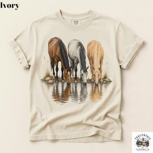 May include: An ivory short-sleeve t-shirt featuring a graphic print of three horses drinking from a body of water. The horses are brown, grey, and tan, with their reflections visible in the water below. The shirt has a crew neckline.