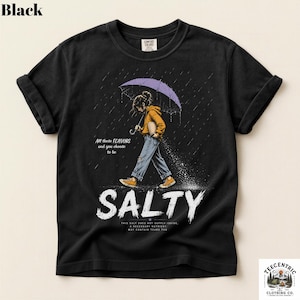 May include: A black t-shirt featuring a graphic of a person in a yellow hoodie and blue jeans walking under a purple umbrella in the rain. The text reads: All these FLAVORS and you choose to be SALTY. THIS SALT DOES NOT SUPPLY IODIDE, A NECESSARY NUTRIENT. MAY CONTAIN TEARS THO.