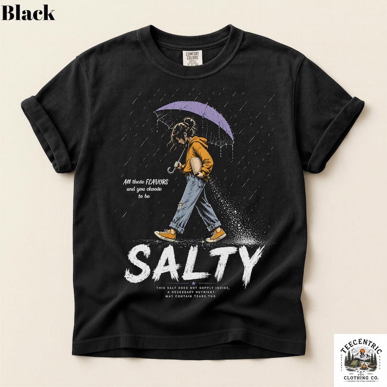 May include: A black t-shirt featuring a graphic of a person in a yellow hoodie and blue jeans walking under a purple umbrella in the rain. The text reads: All these FLAVORS and you choose to be SALTY. THIS SALT DOES NOT SUPPLY IODIDE, A NECESSARY NUTRIENT. MAY CONTAIN TEARS THO.