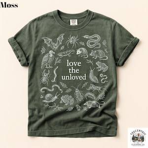May include: A moss green short-sleeve t-shirt featuring a white graphic print of various animals including a bat, spider, snake, and toad surrounding the text "love the unloved" in the center. The shirt has a crew neckline and rolled sleeves.