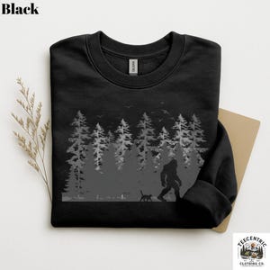 May include: A black crewneck sweatshirt featuring a gray graphic print of a forest with tall pine trees, birds in the sky, and a silhouette of a large creature walking alongside a small cat. The garment has a standard ribbed neckline.