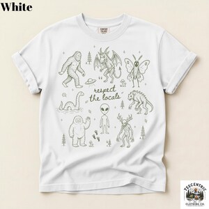 May include: A white short-sleeve t-shirt featuring a dark green line art graphic of various cryptids and the text "respect the locals" in the center. The design includes illustrations of Bigfoot, a mothman, an alien, and other creatures.