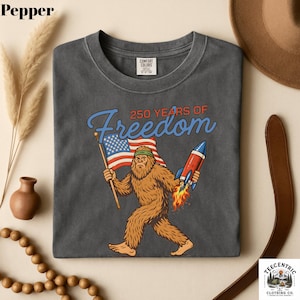 K&ouml;nnte beinhalten: Dunkelgraues T-Shirt mit einem Bigfoot-Motiv, das eine amerikanische Flagge und eine Rakete h&auml;lt, mit dem Text "250 YEARS OF Freedom". Das T-Shirt ist aus einem weichen Material.
