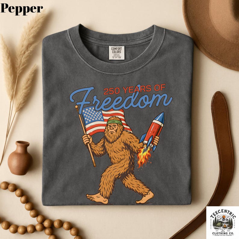K&ouml;nnte beinhalten: Dunkelgraues T-Shirt mit einem Bigfoot-Motiv, das eine amerikanische Flagge und eine Rakete h&auml;lt, mit dem Text "250 YEARS OF Freedom". Das T-Shirt ist aus einem weichen Material.