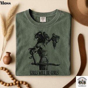 May include: Moss green t-shirt with a graphic of a woman holding a sword and a severed head. The text "GIRLS WILL BE GIRLS" is printed below the graphic. The shirt is made of Comfort Colors material.