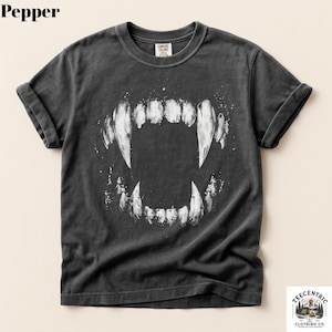 May include: A charcoal gray short-sleeve t-shirt featuring a large, white, graphic print of a set of sharp, bared animal teeth in the center. The shirt has a crew neckline and rolled sleeves, with the word Pepper printed in the top left corner.