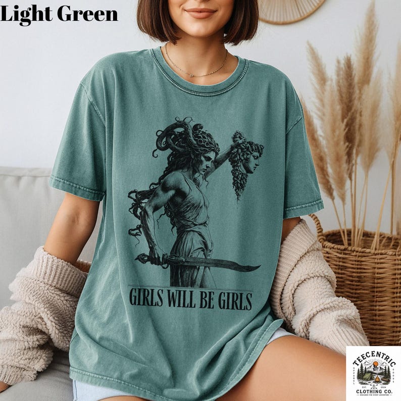 May include: Light green t-shirt with a black graphic of a woman holding a sword and a severed head. The text "GIRLS WILL BE GIRLS" is printed below the graphic. The shirt is from Teecentric Clothing Co.