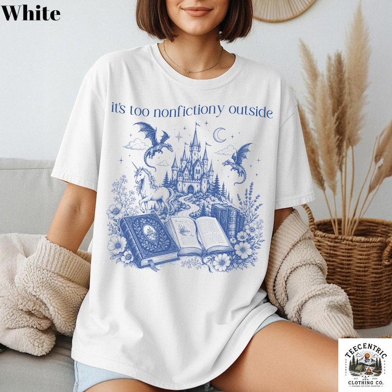 May include: White t-shirt with a blue fantasy graphic. The design features a castle, dragons, a unicorn, and books, with the text "it's too nonfictiony outside". A whimsical design for book lovers and fantasy enthusiasts.