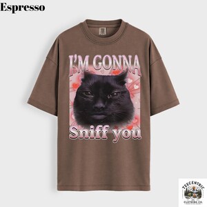May include: Espresso-coloured t-shirt with a black cat graphic and the text "I'M GONNA Sniff you" in pink. The design includes pink hearts and a butterfly. The tee has a crew neck and short sleeves.