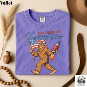 K&ouml;nnte beinhalten: Ein lilafarbenes T-Shirt mit einem Cartoon-Bigfoot, der eine amerikanische Flagge und eine Rakete h&auml;lt, mit dem Text "250 YEARS OF Freedom". Das T-Shirt hat einen Rundhalsausschnitt und ein Comfort Colors-Etikett.
