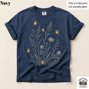 May include: A navy blue t-shirt featuring a white line-art graphic of wildflowers and grasses, accented with yellow firefly illustrations. The text on the shirt reads: Navy, This is a flat print, not actually glow, and TeeCentric Clothing Co.
