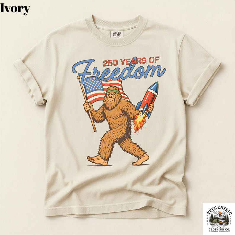 K&ouml;nnte beinhalten: Elfenbeinfarbenes T-Shirt mit einem Bigfoot-Motiv, das eine amerikanische Flagge und eine Rakete tr&auml;gt, mit dem Text "250 Years of Freedom". Das T-Shirt hat hochgekrempelte &Auml;rmel und einen Rundhalsausschnitt.