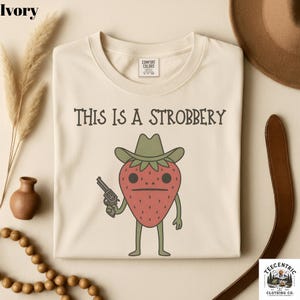 May include: Ivory-colored sweatshirt with the text "THIS IS A STROBBERY" above a cartoon strawberry wearing a cowboy hat and holding a gun. The strawberry is red with green leaves and a sad expression. The shirt has a crew neck.