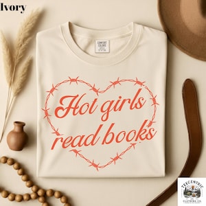 May include: An ivory short-sleeve t-shirt featuring a red graphic of a heart made from barbed wire. Inside the heart, the text reads "Hot girls read books" in a red cursive font. The shirt has a crew neckline and a Comfort Colors brand label.