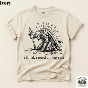 May include: Ivory-colored t-shirt featuring a black illustration of a knight kneeling with a sword, arrows in his back, and the text "i think i need a long rest". The shirt has rolled-up sleeves and a crew neck.