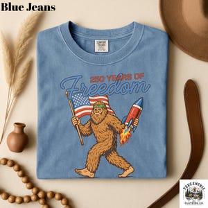 K&ouml;nnte beinhalten: Hellblaues T-Shirt mit einer Grafik von Bigfoot, der eine amerikanische Flagge und eine Rakete h&auml;lt, mit dem Text "250 YEARS OF Freedom". Das T-Shirt ist von Comfort Colors.