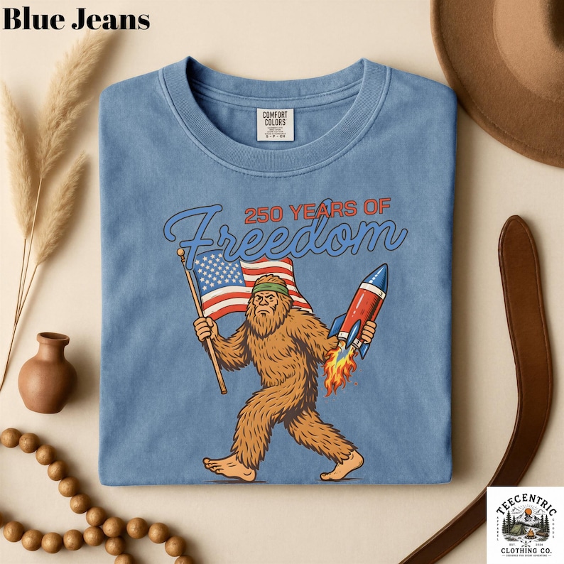 K&ouml;nnte beinhalten: Hellblaues T-Shirt mit einer Grafik von Bigfoot, der eine amerikanische Flagge und eine Rakete h&auml;lt, mit dem Text "250 YEARS OF Freedom". Das T-Shirt ist von Comfort Colors.