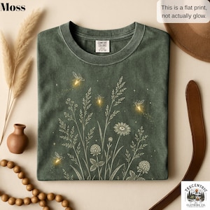 May include: A moss green t-shirt featuring a white line art graphic of wildflowers and grasses with glowing yellow fireflies and small stars. The shirt has a crew neckline and a Comfort Colors brand label. Text on the image reads: This is a flat print, not actually glow.