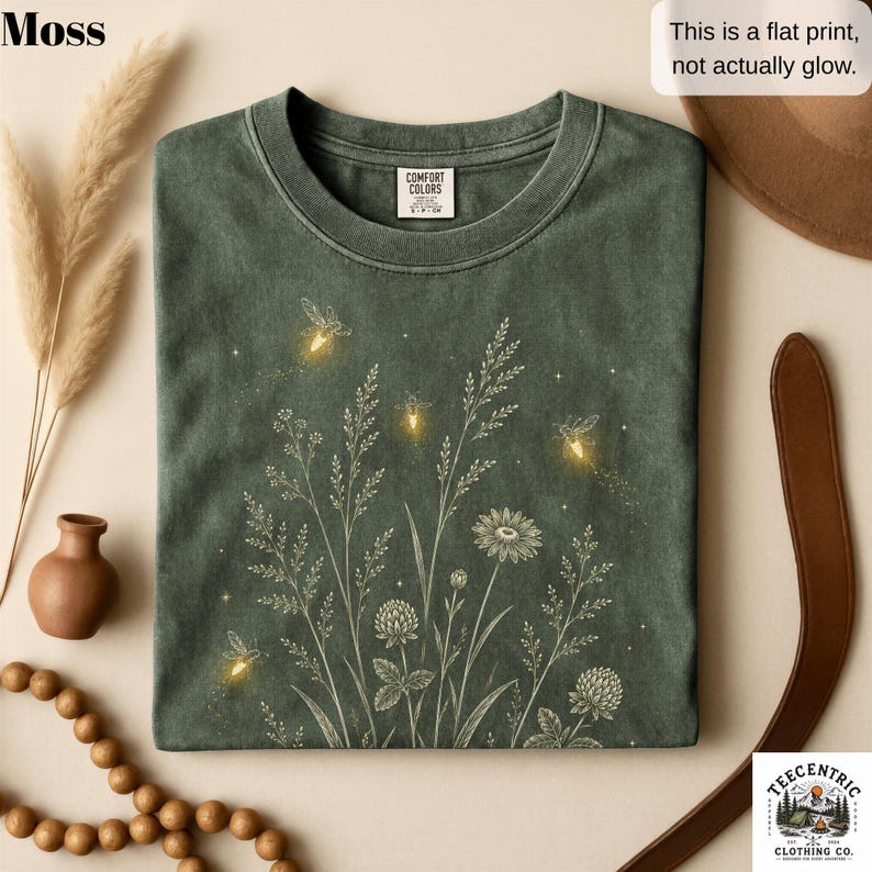 May include: A moss green t-shirt featuring a white line art graphic of wildflowers and grasses with glowing yellow fireflies and small stars. The shirt has a crew neckline and a Comfort Colors brand label. Text on the image reads: This is a flat print, not actually glow.