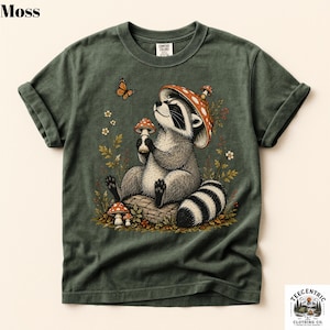 May include: A moss green short-sleeve t-shirt featuring a graphic of a raccoon sitting on a log, wearing a red and white spotted mushroom cap as a hat. The raccoon holds a small mushroom, with a butterfly and wildflowers surrounding it.