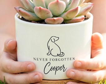 May include: A white ceramic cylindrical planter containing a green and pink succulent. The planter features a black line drawing of a dog and the text "NEVER FORGOTTEN Cooper" in black ink. The pot sits on a circular light brown wooden base.