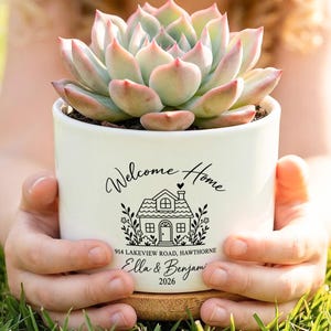 Può includere: Un vaso in ceramica bianca contenente una pianta succulenta. Il vaso presenta una scritta nera che recita Welcome Home, 914 LAKEVIEW ROAD, HAWTHORNE, Ella & Benjamin e 2026, insieme a un'illustrazione grafica di una casa e motivi floreali.