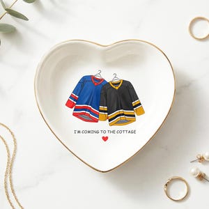 May include: Heart-shaped ceramic trinket dish with a gold rim, featuring illustrations of two hockey jerseys, one blue and red, the other black and gold. Text reads "I'M COMING TO THE COTTAGE" with a red heart. Decorative jewelry surrounds the dish.