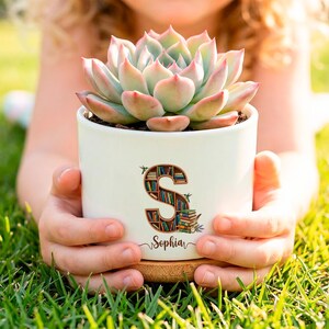 Peut inclure: Un pot de fleurs en céramique blanche contenant une plante succulente verte et rose. Le pot présente une grande lettre S remplie d'une illustration de bibliothèque colorée et le prénom Sophia écrit en lettres cursives noires.