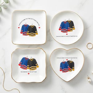 May include: Four white ceramic trinket dishes with gold rims. Each dish features a hockey jersey design in blue, black, and yellow, with the text "I WOULD COME TO THE COTTAGE FOR YOU" or "I'M COMING TO THE COTTAGE".