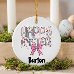 May include: A white, round Easter ornament with the words "HAPPY EASTER" in pastel colors and a pink bow. The name "Burton" is printed in black below the bow. The ornament hangs from a gold string. Easter eggs and candles are in the background.